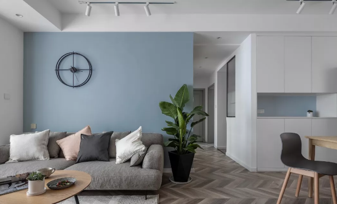 灰色布艺沙发点缀几个低饱和色调的靠枕装饰，在纯粹的蓝灰色沙发背景上装饰造型挂钟，一角的望鹤兰增添生机